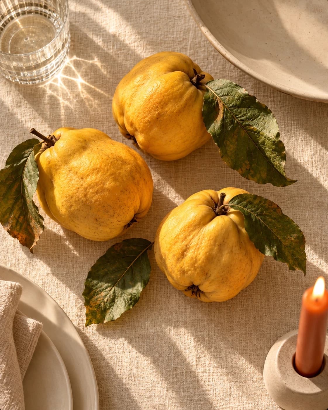 Three ripe quinces with their leaves on a linen tablecloth in late afternoon sun, beside a glass of water and a lit candle.