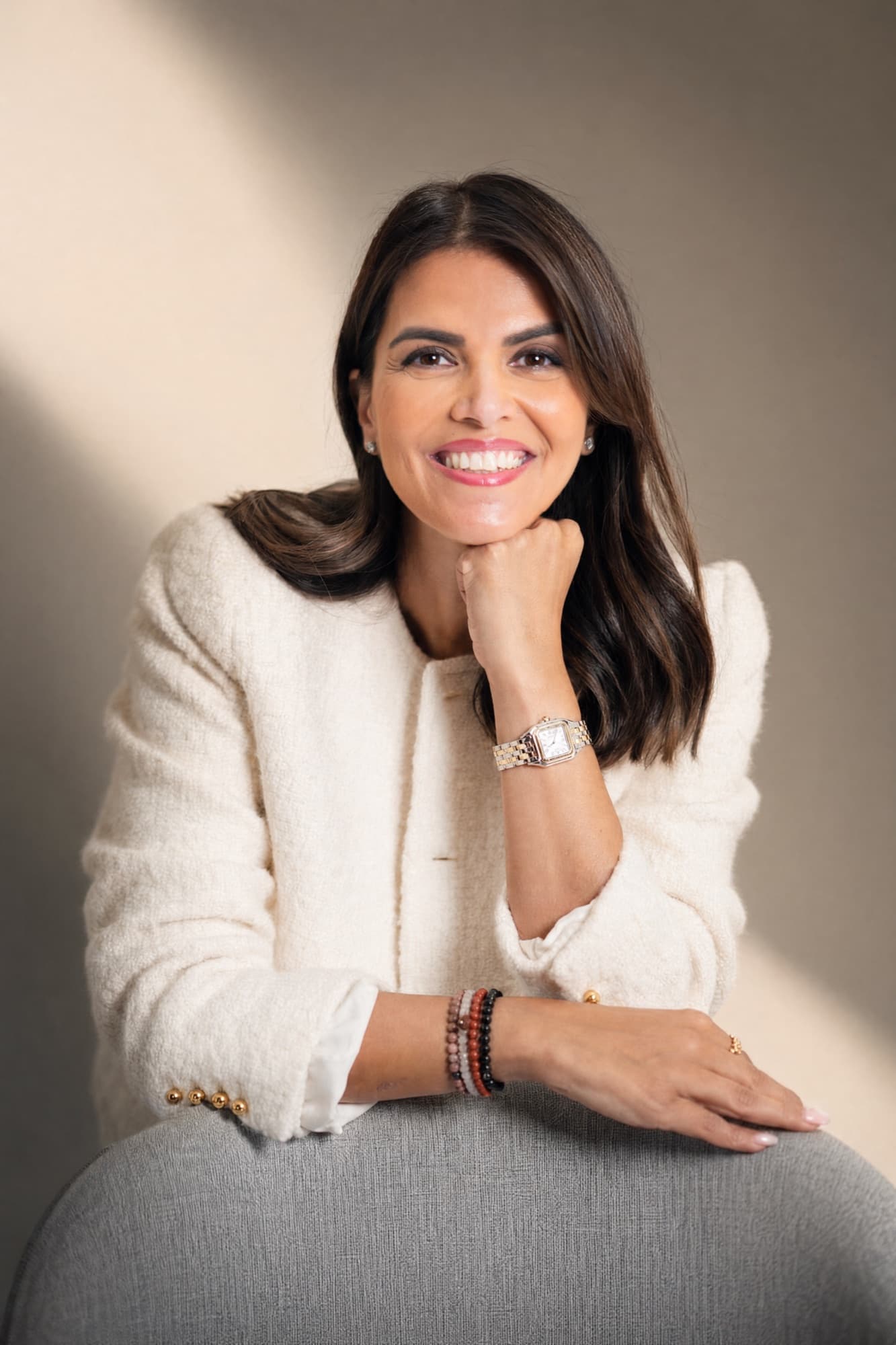 Zahra Abdalla smiling, resting her chin on her hand, in a cream jacket against a warm, sunlit wall.