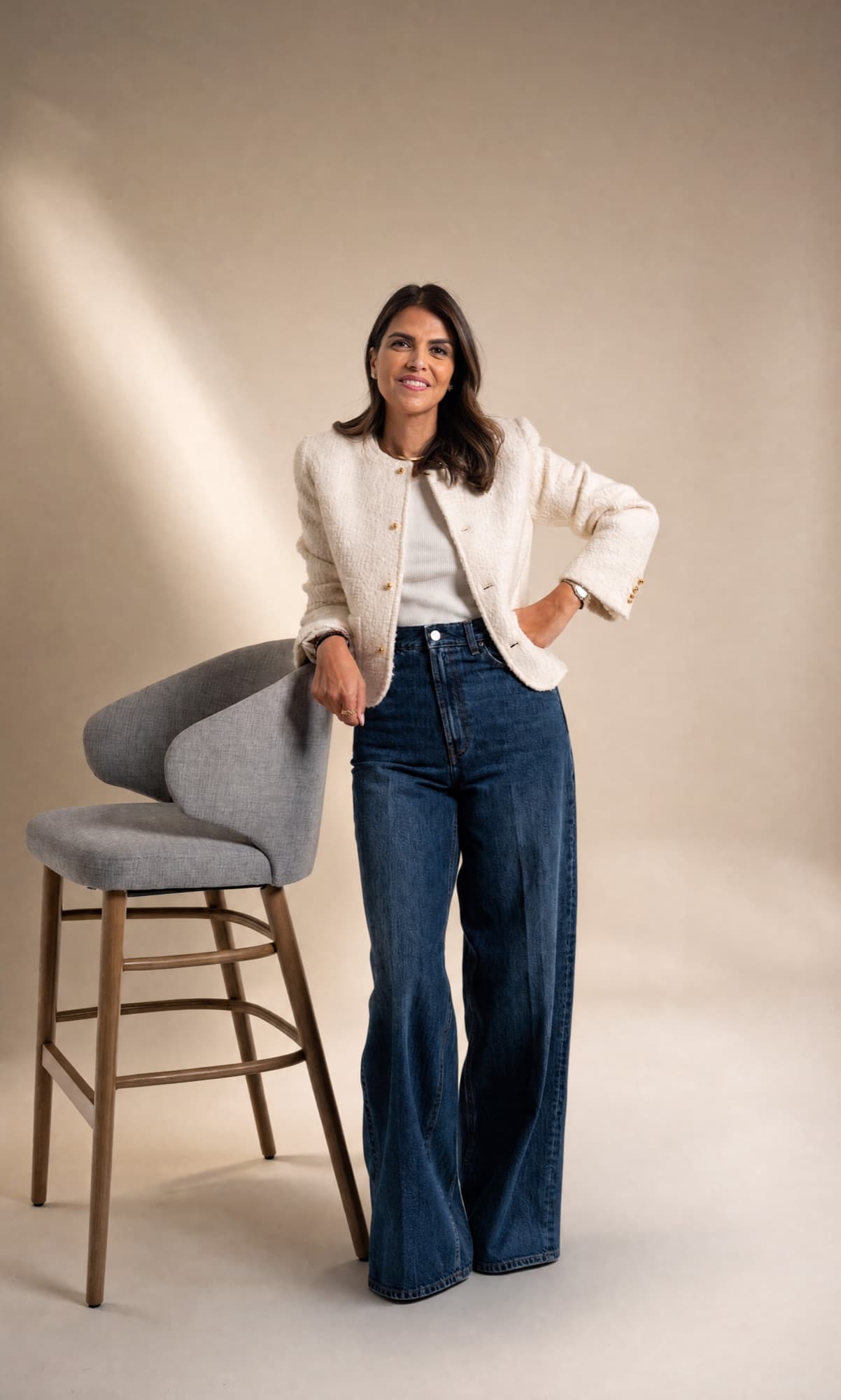 Zahra Abdalla standing beside a grey stool in a cream jacket and wide-leg jeans.
